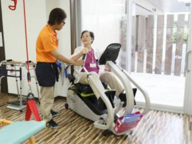 リハビリ機器と利用者 トレーニングコーナー 笑顔で接するスタッフと安心してトレーニングに臨む利用者の姿。快適な環境でのサポートがここにはあります。