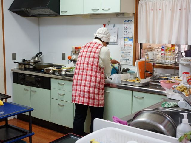 調理中のキッチン風景 食堂スタッフ 明るく清潔なキッチンで、一人のスタッフが丁寧に食事の準備をしています。手作りの温もりある料理をご提供しております。