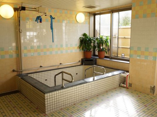 浴室の大きな浴槽には、体を支える介護用機器が設置されている。床と壁はタイル貼りになっており、入りやすいよう手すりが付いている。