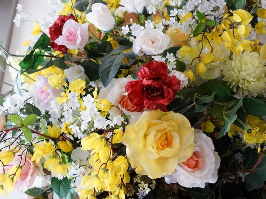 装飾用の造花である。赤、ピンク、黄色などのバラがたくさんあり、細部まできれいに作られている。葉っぱや小さな花も飾られている。
