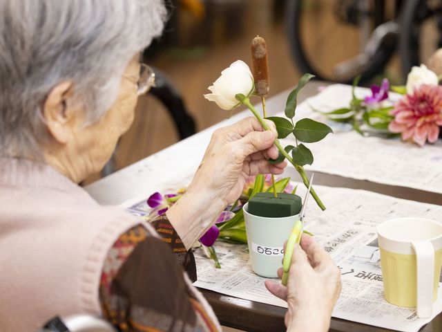 生け花を楽しむ利用者 多目的室   明るいテーブルで生け花を楽しむ様子です。手元で花を整えながら、落ち着いて過ごせる場になっています。