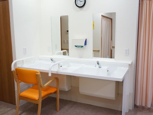 洗面台が2つ、その上に鏡とペーパータオルが設置されている。時計もある。洗面台には手摺があり、椅子が1脚置かれている。