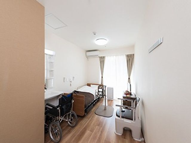 車椅子のある居室 居室   明るい居室にベッドと車椅子が置かれ、窓辺からやわらかな光が入っています。介護の準備や動線を確認しやすい、すっきりした空間です。