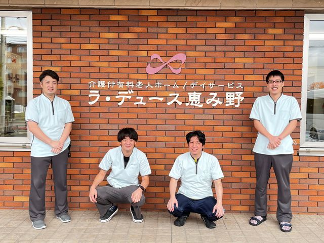 スタッフイメージ スタッフ 壁に掲げられた看板の前に立つ笑顔のスタッフたち。チームワークの良さが感じられる様子です。