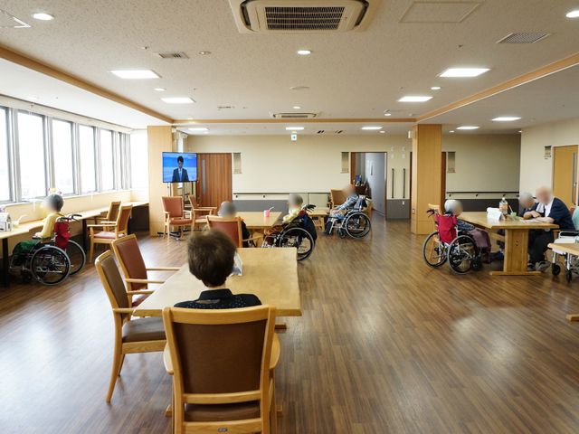 多機能空間と利用者 共有リビング ゆったりとした空間で利用者がそれぞれの時間を過ごしています。木目調の家具が温かみのある雰囲気を演出しています。