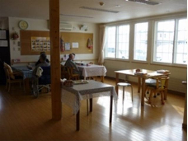 共有スペースの明るい居室 共有スペース ゆったりとした空間で利用者同士が食事をとる様子が伝わります。大きな窓からは自然光が差し込み、温かい雰囲気を感じさせる設計です。