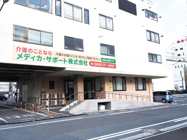 白い建物のエントランス 外観 清潔感がある白い外壁に、緑色の看板が目を引く建物です。出入りしやすいように、斜めのアプローチが整備されております。