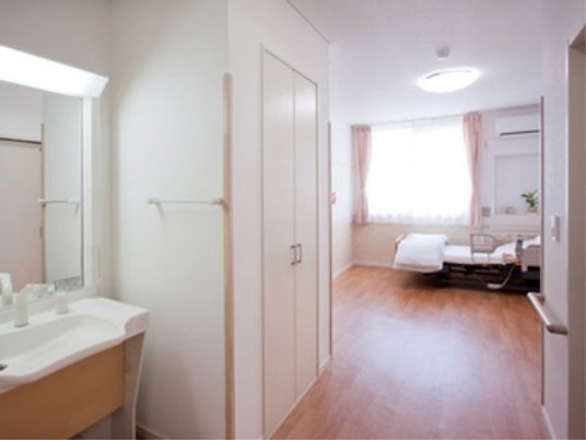 施設の写真 木目調のフローリングに白を基調とした居室には、洗面所や備え付けクローゼットが完備され、介護用ベッドが設置されている。