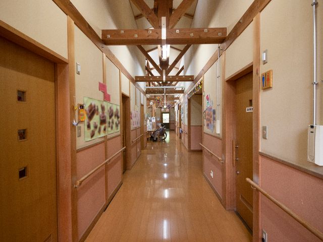 廊下に人と手すり 廊下 温かみのある木造廊下で、手すりが整備され安全な移動が考慮されています。壁には掲示物や飾りがあり、訪れる人を和ませる工夫が見られます。