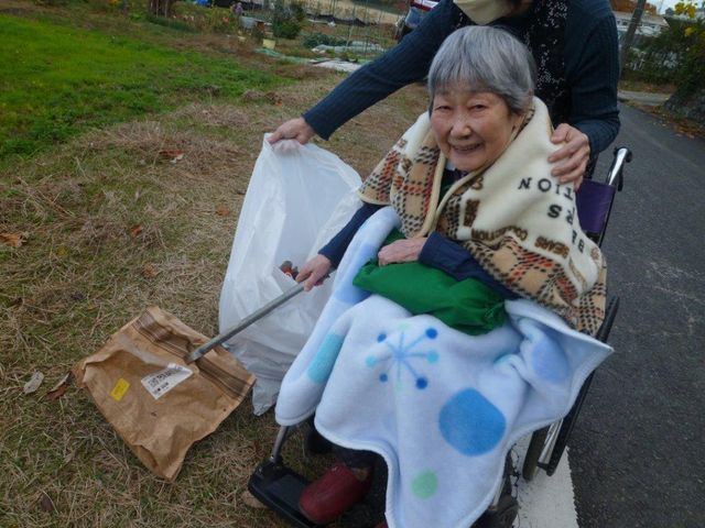 車椅子の高齢者と介助者 ゴミ拾い お手伝いを受けながら外の空気を感じている利用者の笑顔が心温まります。ケアの仕事には人とのふれあいがあります。