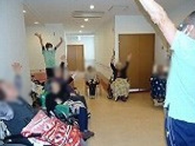 体操する利用者たちの光景 廊下 広々とした廊下で、数名の方が座りながら元気に体操しています。明るい雰囲気の中で、楽しく体を動かすことができます。