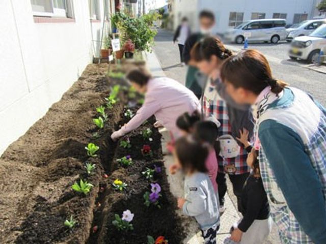 園芸活動を眺める様子 庭 庭で花を植えながら入居者とスタッフが楽しい時間を過ごしています。子どもたちも参加しています。