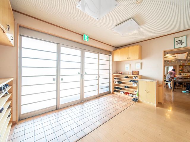 エントランスの和やかな光景 玄関 ゆとりあるエントランスでは、履物が整然と並び、奥には人々が暮らしを楽しむ姿が見えます。光の差し込む心地良い空間で、新たな職場をご検討ください。