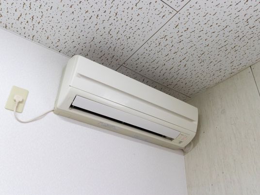 白い壁の天井近くにエアコンが取り付けられ、左側に向かってコンセントが伸びている。天井には模様が付いている。