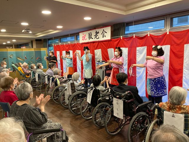 イベント風景の活動室 食事 スタッフが手作りの太鼓演奏を披露し、車椅子の方々が手拍子で盛り上がっています。明るい笑顔で交流する姿が印象的な一コマです。