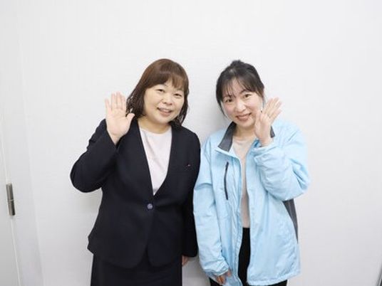 笑顔で手を振る2人の人物が並ぶ室内  