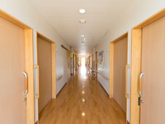 施設の写真 施設内の廊下は、白色と茶色で統一したシンプルな内装となっている。両サイドには、大きな引戸の取りつけられた居室がたくさん並んでいる。