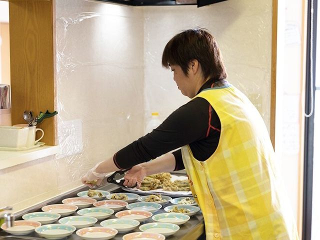 食事準備をするスタッフ 日常風景 配色の明るいエプロンを着けたスタッフが、丁寧に食事の準備を行っています。温もりのある食事の時間をサポートする仕事に携われます。