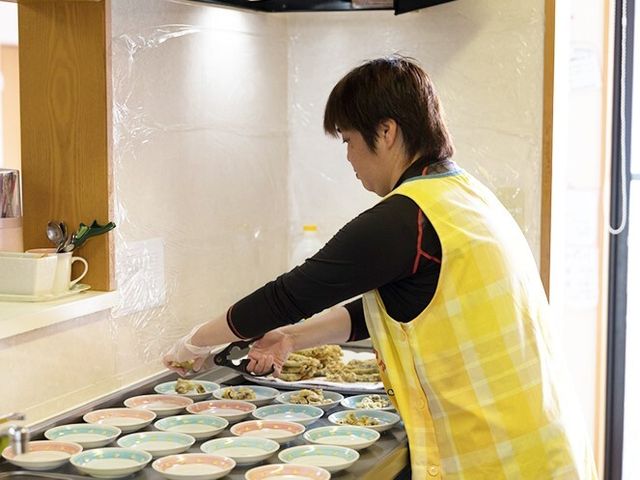 食事準備をするスタッフ 日常風景 配色の明るいエプロンを着けたスタッフが、丁寧に食事の準備を行っています。温もりのある食事の時間をサポートする仕事に携われます。