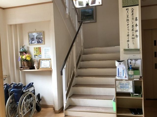 階段と掲示物の廊下 玄関 ゆったりとした空間の廊下には、様々な掲示物があり、情報の共有に配慮されています。車椅子も置かれており、利用者の利便性が考えられています。