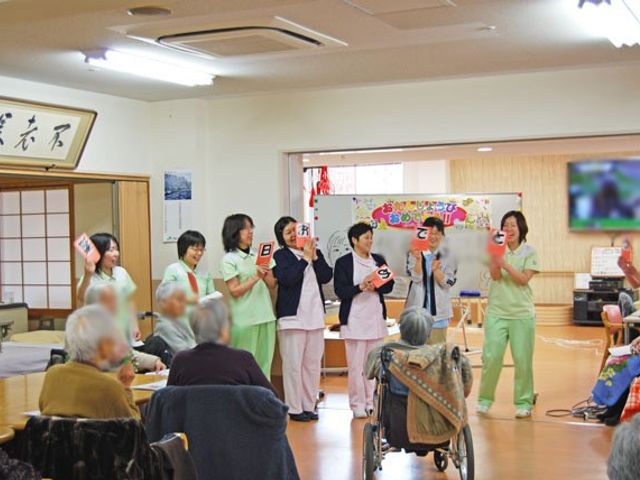 にぎやかな集団の様子 多目的室 広々とした明るい空間で、スタッフと入居者が和やかにコミュニケーションを楽しむ場面です。