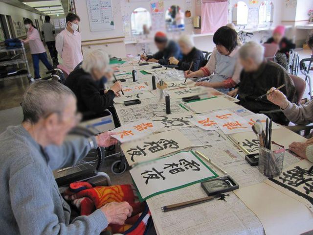 書道を楽しむ高齢者たちの居室 居室 広々とした明るい場所で、皆さんが書道を楽しんでいます。和やかな交流が見られます。