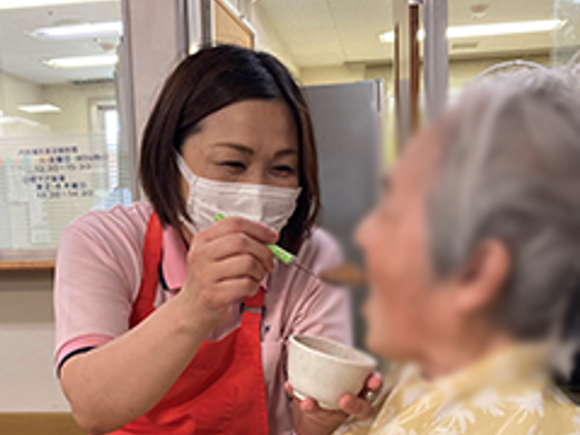 親身な介助の場面 スタッフイメージ 職員が笑顔で利用者に食事を介助しており、親しみやすい雰囲気の中で丁寧なケアが行われています。