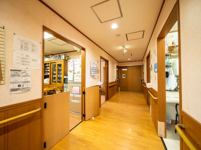 廊下の視点から見た風景 廊下 手すりを備えた落ち着いた色合いの廊下が、利用者の安全な移動をサポートしています。壁には掲示物があり、情報共有のスペースとなっています。