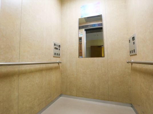 エレベーターの壁には手すりと操作盤があり、鏡が１枚取り付けられている。床は灰色で壁は黄土色になっている。