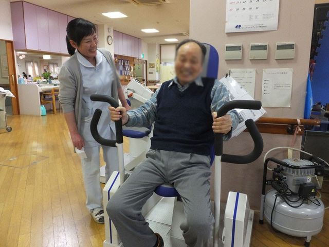 笑顔で運動する場面 機能訓練室 笑顔で機器を使った運動をしており、和やかにサポートするスタッフの姿が見られます。施設の活気を感じる空間です。