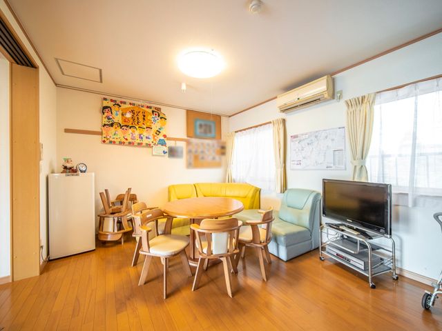 明るいリビングの様子 談話室 木目調の床に輝くリビングは、ゆとりある空間で日々の憩いの場を提供しています。ソファやテレビがあり、のんびりとくつろげる環境です。