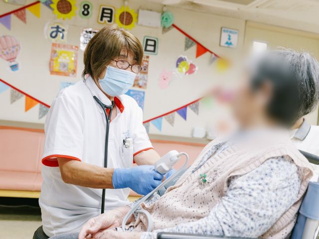 お部屋の診察の様子 スタッフイメージ 医療スタッフが利用者に体調確認を行うシーンです。安心して暮らせる環境が整っています。