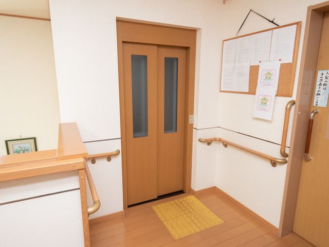 廊下と部屋の扉 エレベーター 壁には掲示物があり、手すり付きの廊下は安全な移動をサポートしています。扉のデザインもシンプルで開閉がしやすそうです。
