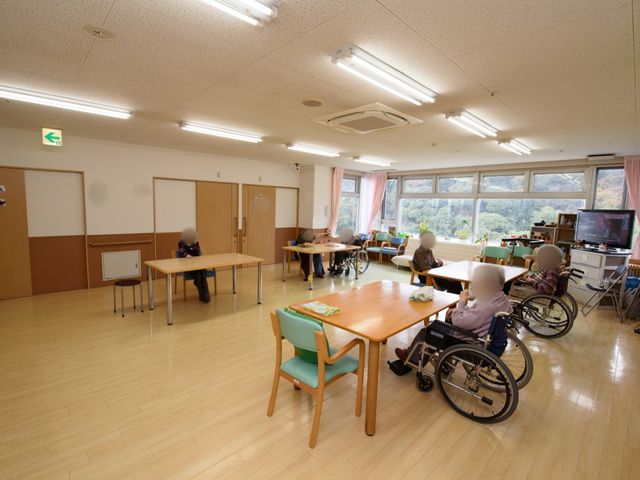 共有スペースの居心地の良い様子 グループホーム 広々としたラウンジでは、ご利用者がくつろいでお話ししたり、テレビを見たりしている場面です。窓からは自然の光が差し込み、温かな雰囲気が漂っています。