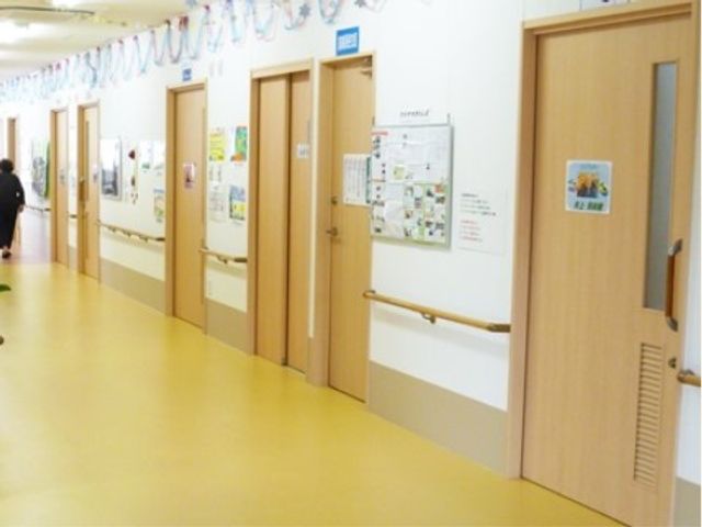明るい廊下と部屋の扉 通路 手すり付きの広々とした廊下は、安心して歩行できる設計です。壁に掲示された情報も目を引き、和やかな雰囲気を演出しています。