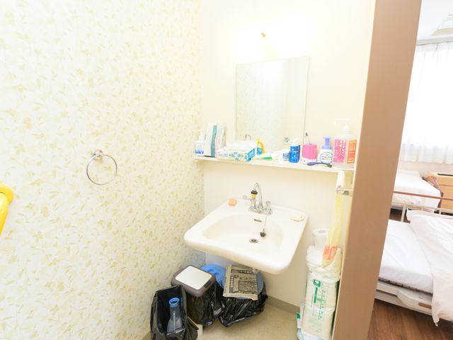 居室の洗面所 洗面台 居室に備え付けられた洗面台は、日常生活に必要な備品を置くためのスペースも確保されています。利用者様が快適にご使用いただける様子が伺えます。