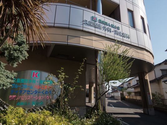 建物の横側には、植栽を施し、シルバーの楕円の看板を設けている。敷地内には、駐車場を備え、アスファルトの上には矢印がある。