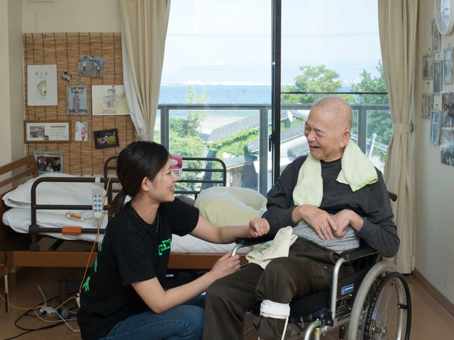 ケアと笑顔の居室風景 明るい窓辺で、スタッフが笑顔で話しをする様子。窓の外には海が広がり、心地よい風景を楽しめます。