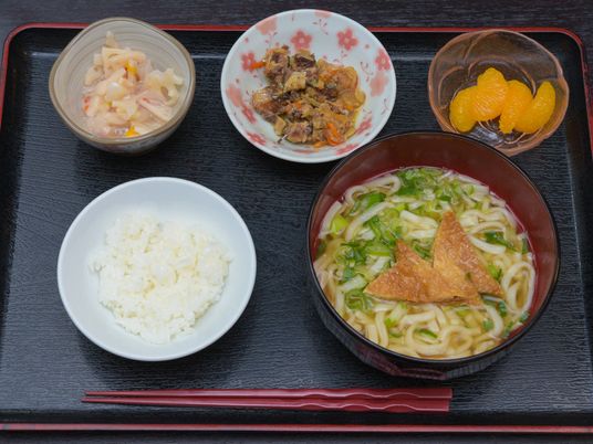 施設の食事は和食を中心に麺類などもある。ご飯、うどん、煮物、サラダ、みかんの缶詰が配膳されているのが写っている。