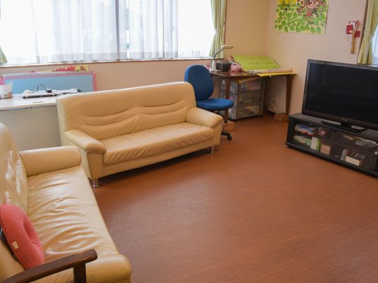 リビングはフローリングの洋室。黄色のソファーが2つ置かれていて、その前にはワイドテレビが設置されている。壁際にはデスクや棚も見える。