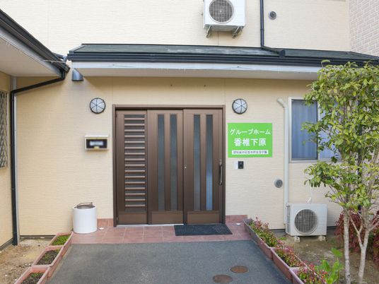 施設の玄関はクリーム色の壁に緑の看板に白で施設名が大きく書かれている。玄関の扉は横開きで扉の横にはインターホンが設置されている。