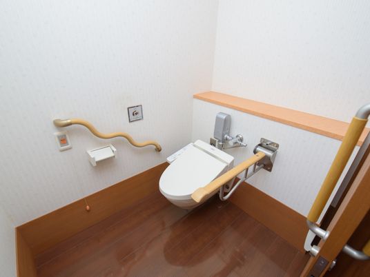 トイレの床は焦げ茶色のフローリングになっている。壁には波型の手すりやトイレットペーパーホルダー、呼び出しボタンが設置されている。