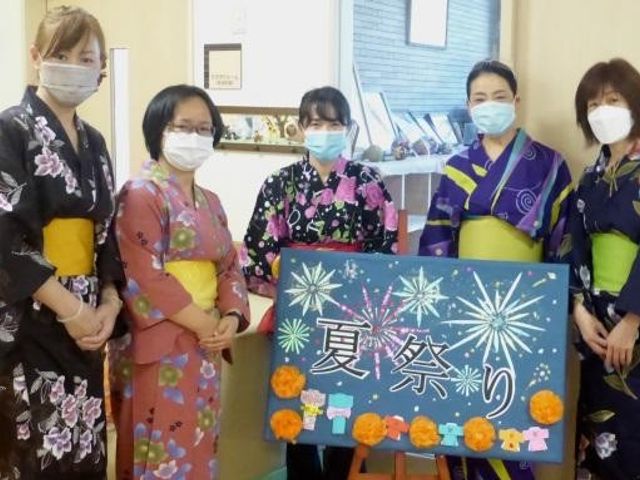 和服姿のスタッフと掲示物 スタッフイメージ 和服を着た笑顔のスタッフが、手作りの掲示物を前に立っています。彩り鮮やかな雰囲気が感じられます。