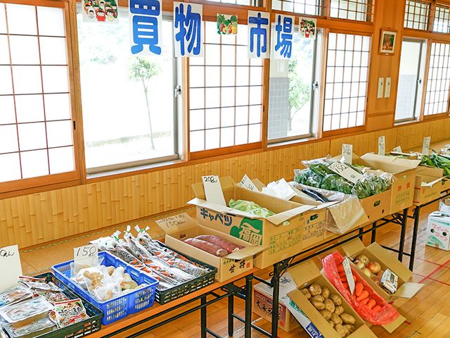 野菜販売テーブル 多目的室 自然光が差し込む空間で、多様な商品が並ぶ買い物イベントが開催されています。