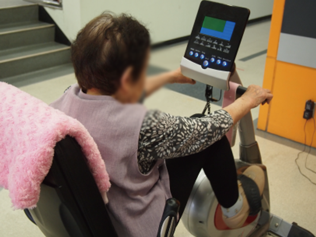 機能訓練中の様子 機能訓練室 利用者が専用機器で運動しながら、日々健康維持に取り組んでいます。女性が汗を流す姿が印象的です。