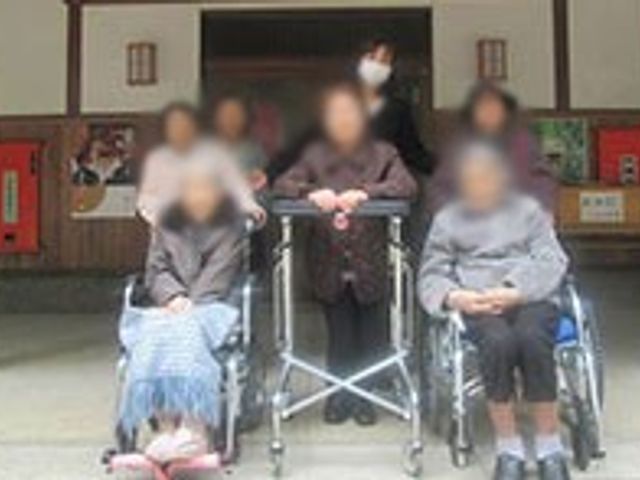 車椅子のある集合写真 外観 建物前で集まる利用者とスタッフが和やかに交流しています。リラックスした雰囲気です。