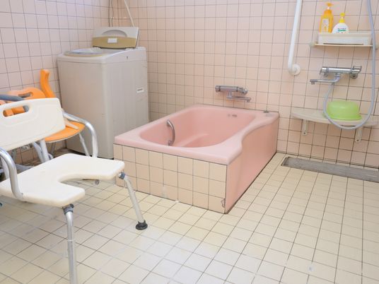 施設の写真 淡いピンク色のタイルが貼られた浴室がある。洗い場にはシャワー椅子があり、壁や浴槽には手すりがある。