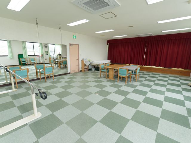 広々とした機能訓練室 共有スペース 緑とベージュのチェック柄の床が印象的な訓練スペースです。明るくて開放感のある空間でスタッフとともにリハビリに取り組むことができます。