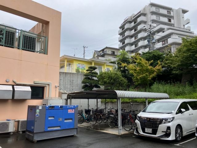 建物前の駐車場 外観   建物前に駐車スペースがあり、屋根付きの駐輪場も見えます。周囲に緑があり、落ち着いた雰囲気です。