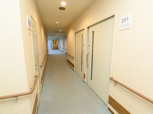 施設の写真 廊下には居室やトイレのドアがあり、白い表札が出ている。天井のライトは、丸型で中央に等間隔に並んでいる。